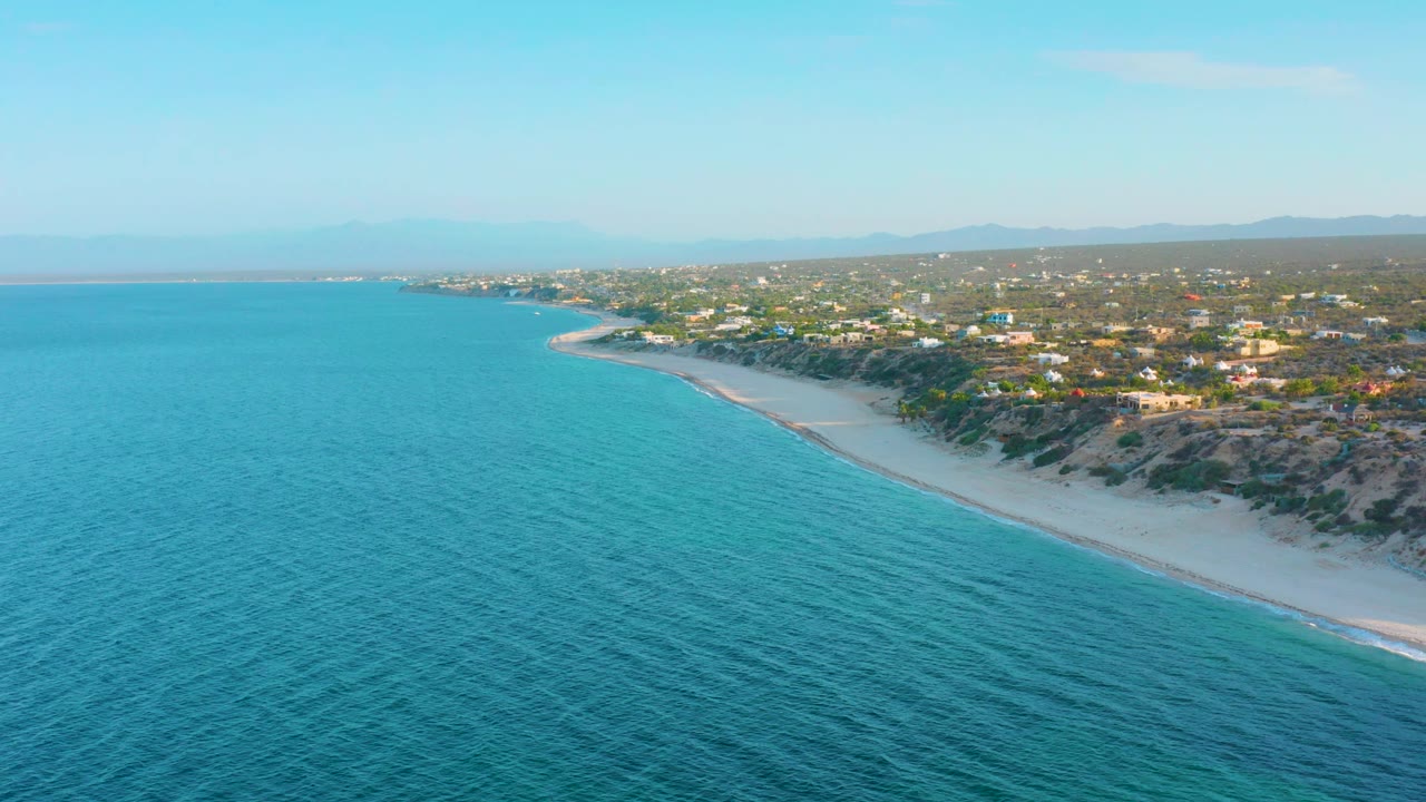 Aerial coastline with turquoise water, sandy beach, scrub dunes, and small buildings inland