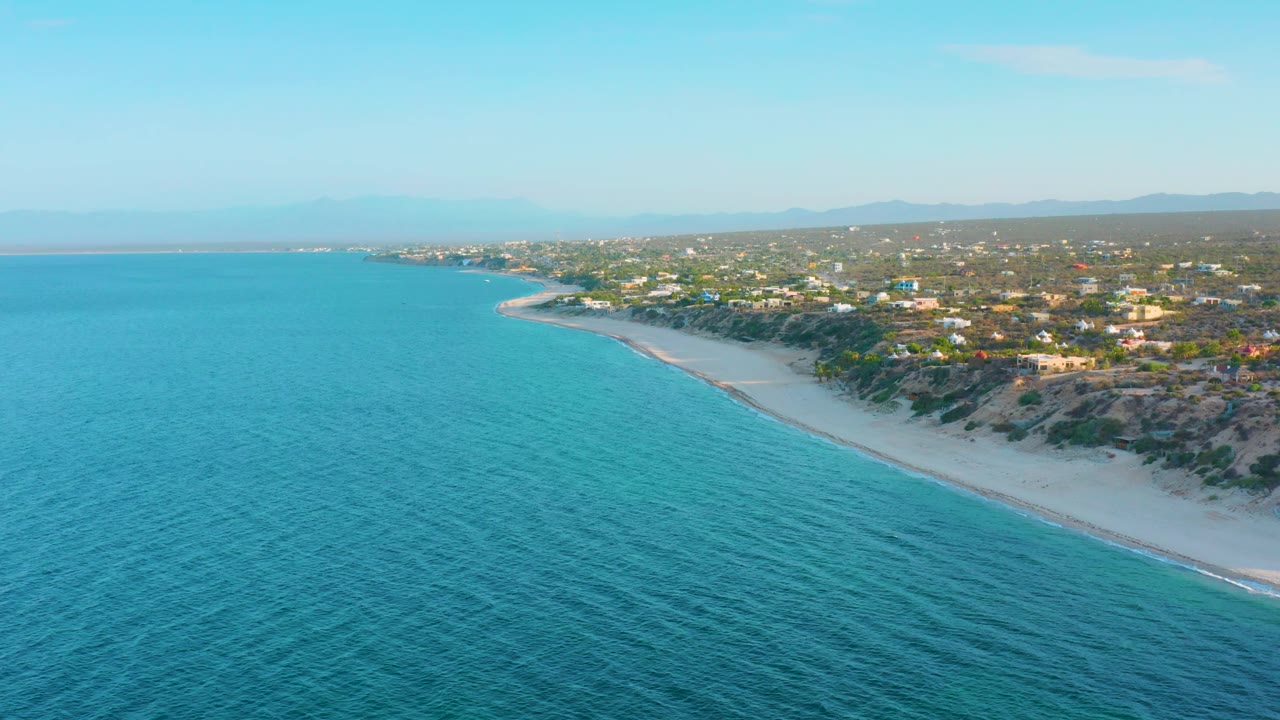 Wide aerial frame over El Sargento La Ventana, Mexico, showing rippled blue water, the pale beach running diagonally, dark coastal vegetation, low buildings inland, distant hills, and no visible people.