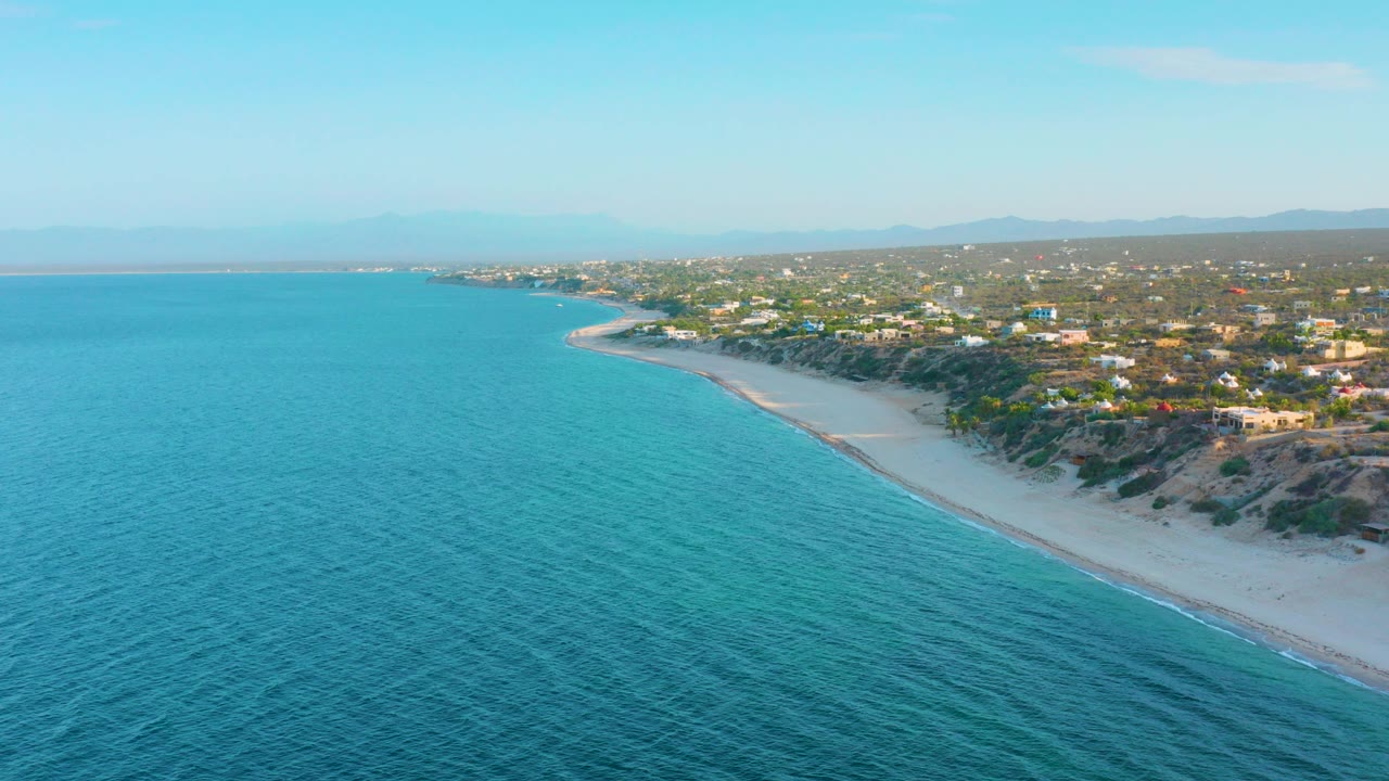 Aerial view over El Sargento La Ventana, Mexico, with turquoise water in the foreground, a pale beach curving right, inland homes beyond the bluffs, distant hills, and no visible people.