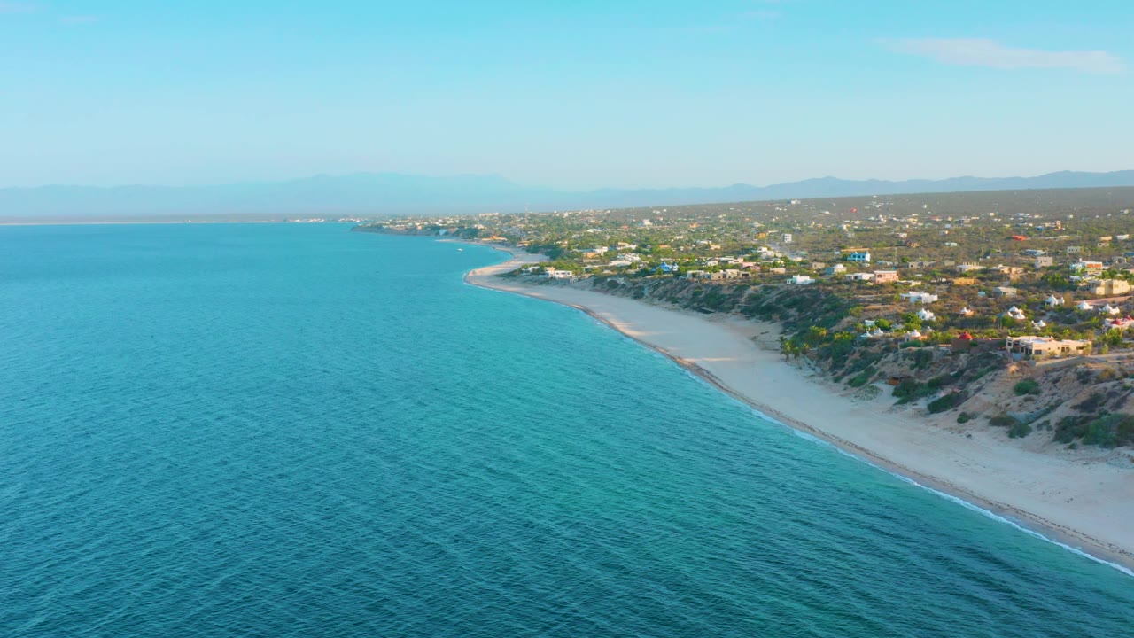 Wide aerial view of El Sargento La Ventana, Mexico, with open turquoise sea on the left, a diagonal sandy shoreline on the right, low scrub-covered bluffs, inland homes, distant hills, and no visible people.
