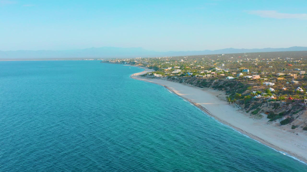 Opening aerial frame over El Sargento La Ventana, Mexico, with turquoise water in the foreground, a pale curving beach, coastal homes on the bluff, distant mountains, and no visible people.