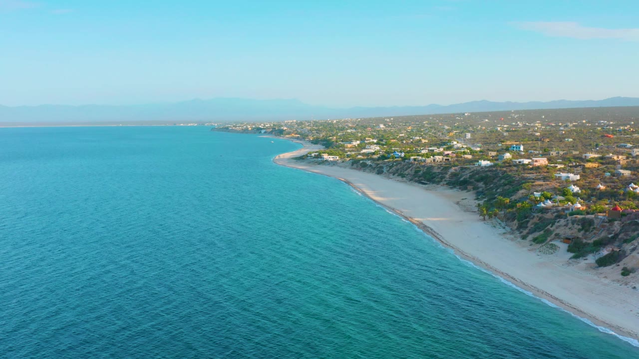 The aerial composition in El Sargento La Ventana, Mexico, shows open turquoise water dominating the left side, beach and bluff on the right, a coastal settlement beyond, and no visible people.