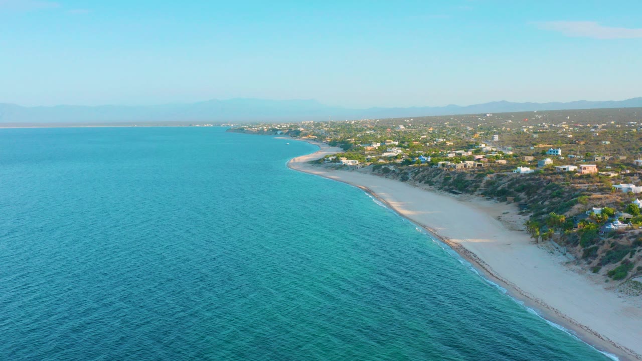 At 0:00, an aerial view over El Sargento La Ventana shows turquoise water, a curved white sand beach, scrub-covered land, distant mountains, and no visible people.