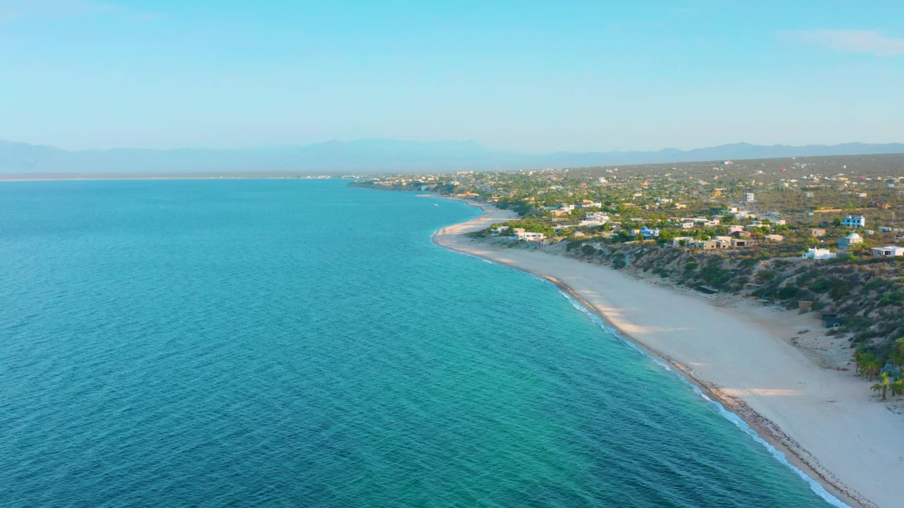 Aerial view over El Sargento La Ventana, Mexico, with blue ocean dominating the foreground, the sandy shoreline bending right, coastal homes inland, distant hills, and no visible people.