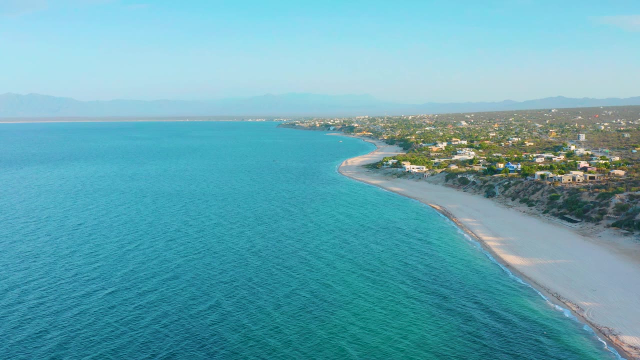 Wide aerial coastline with turquoise water, sandy beach, houses, and hills.