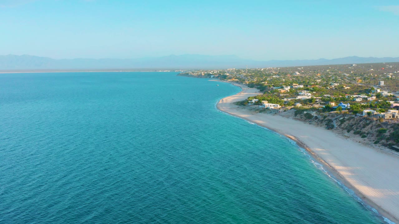 Opening aerial frame over El Sargento La Ventana, Mexico, with turquoise water in the foreground, a pale beach curving right, low coastal buildings inland, hazy mountains in the distance, and no visible people.