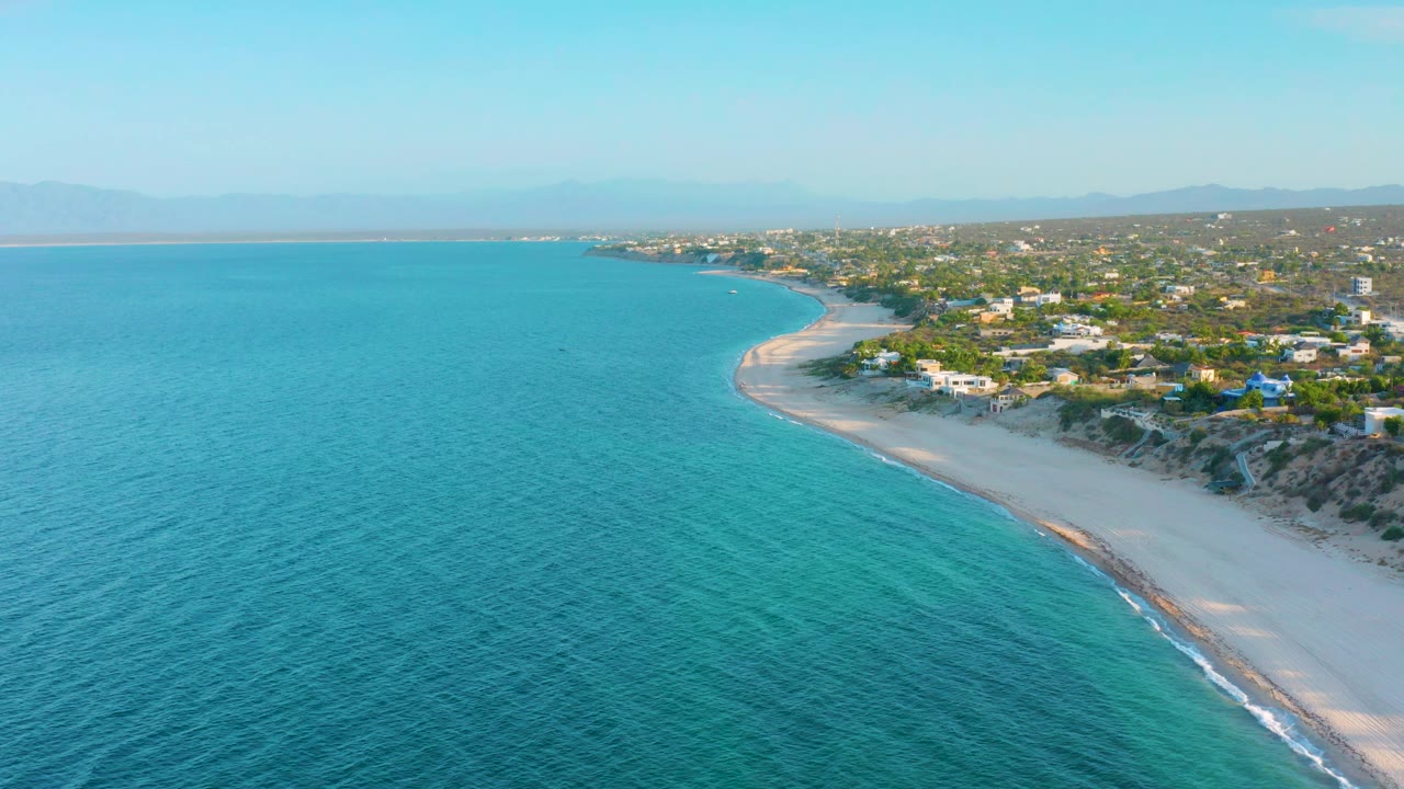Wide aerial coastline with blue-green ocean, pale beach, dune edge, homes, and hazy mountains.