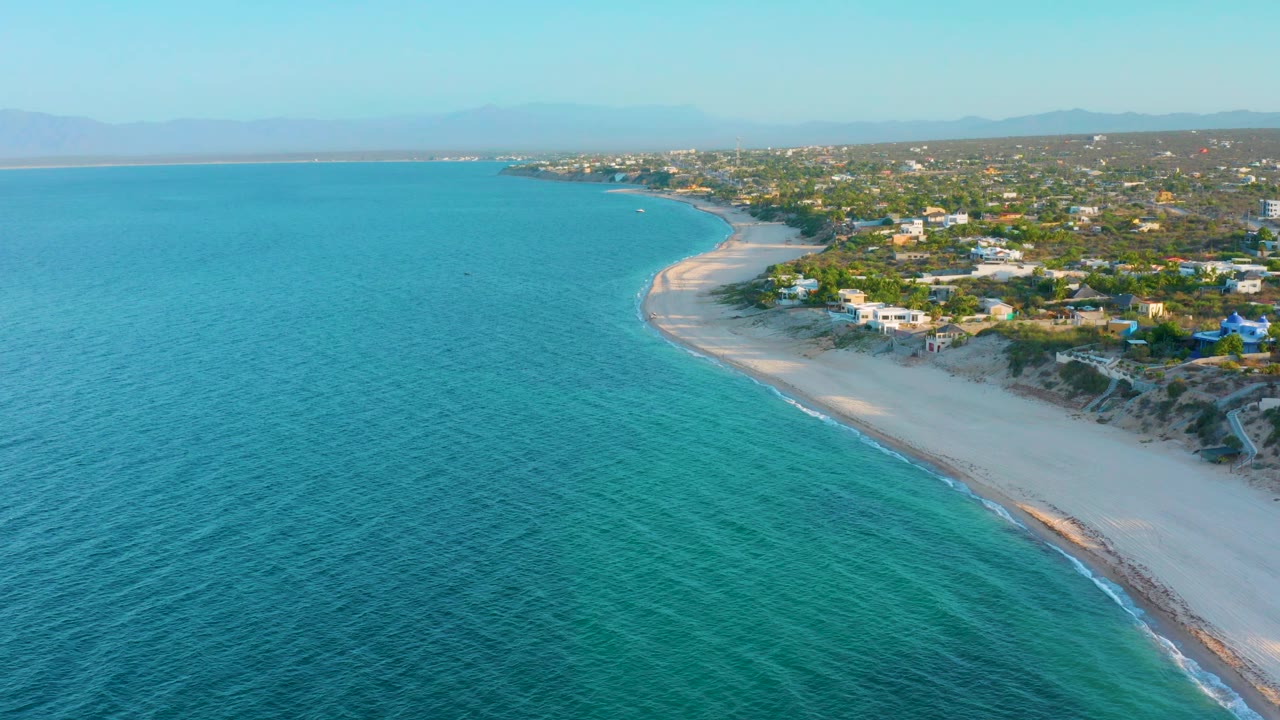 Wide drone view of clear green-blue sea along a pale beach with homes inland.