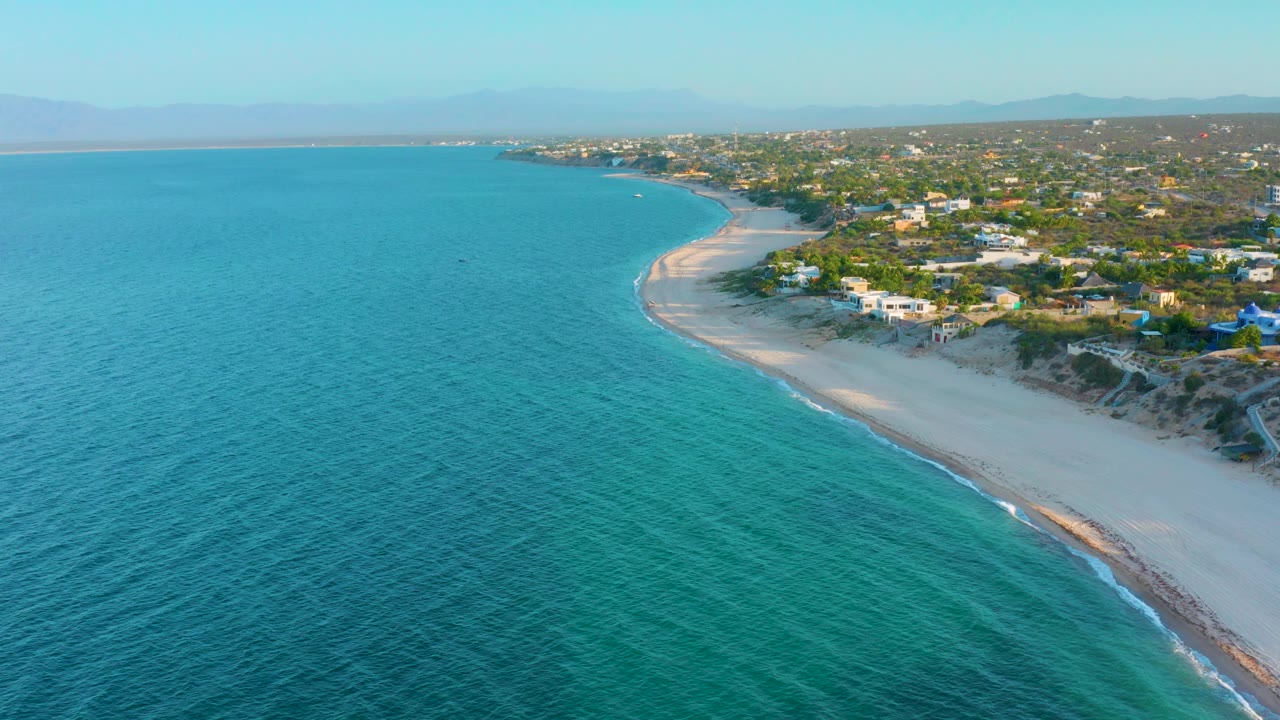 At 0:02, textured turquoise water occupies most of the frame while the sandy El Sargento La Ventana beach arcs along the right edge below scattered homes, with no visible people.