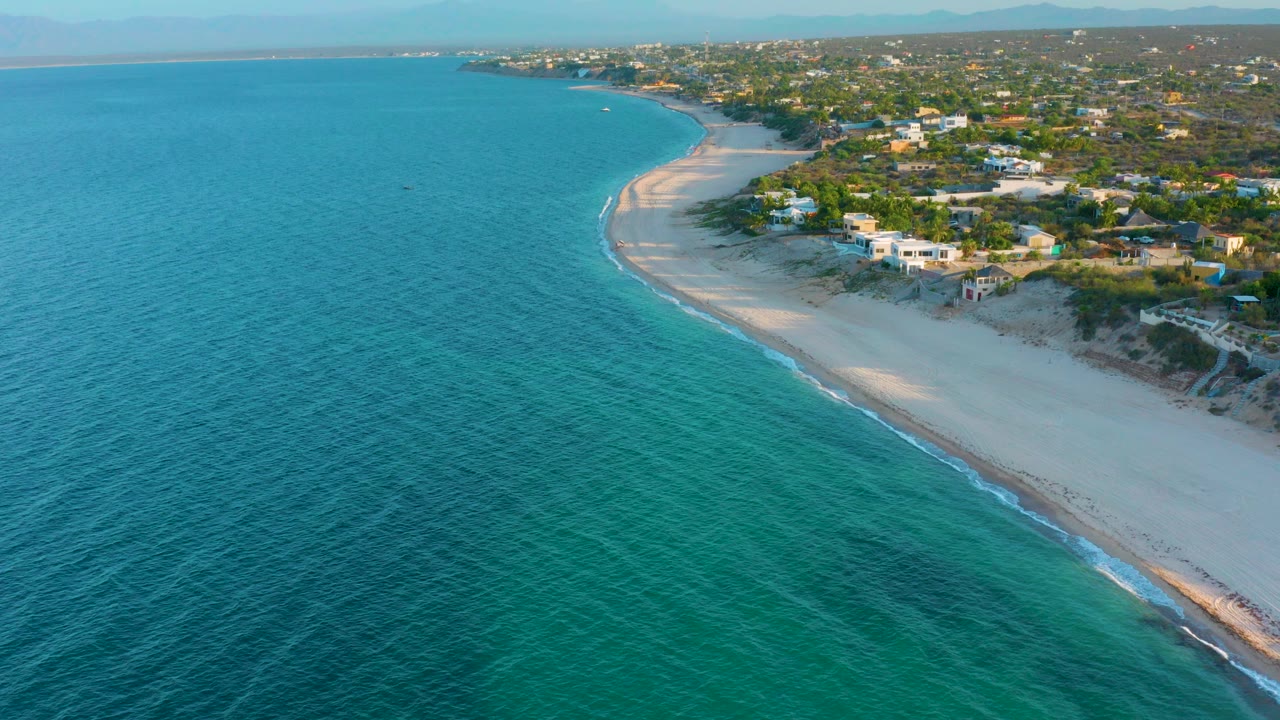 Opening aerial view in El Sargento La Ventana, Mexico, with turquoise water filling the foreground, a pale beach curving to the right, beach houses beyond the dunes, and no visible people.