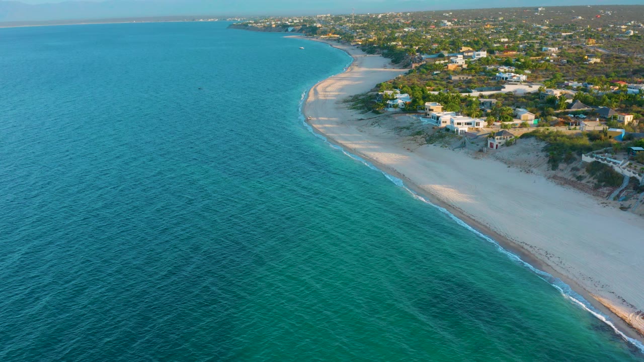 High coastal view in El Sargento La Ventana, Mexico, with more open water across the foreground, the beach running diagonally along the right edge, low houses above the sand, and no visible people.