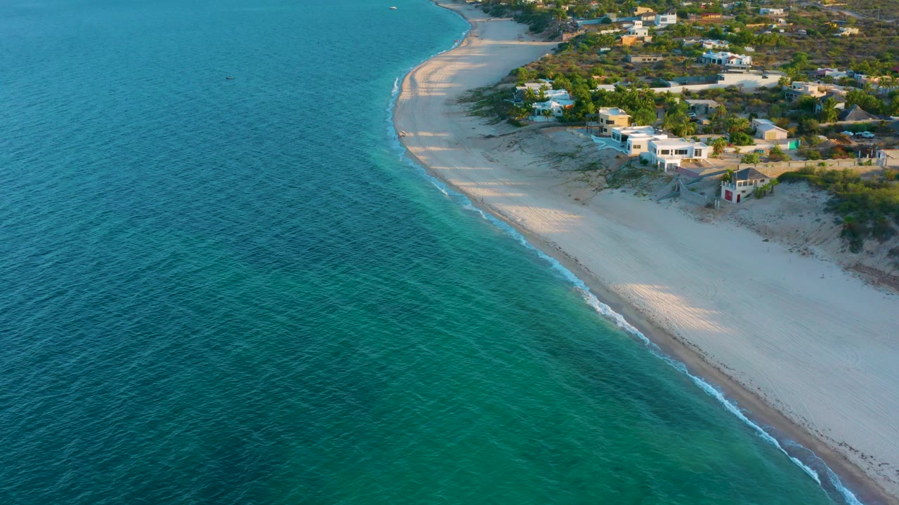 Drone-height view of El Sargento La Ventana, Mexico with the shoreline running diagonally, darker patches under the water, low homes above the sand, and no visible people.