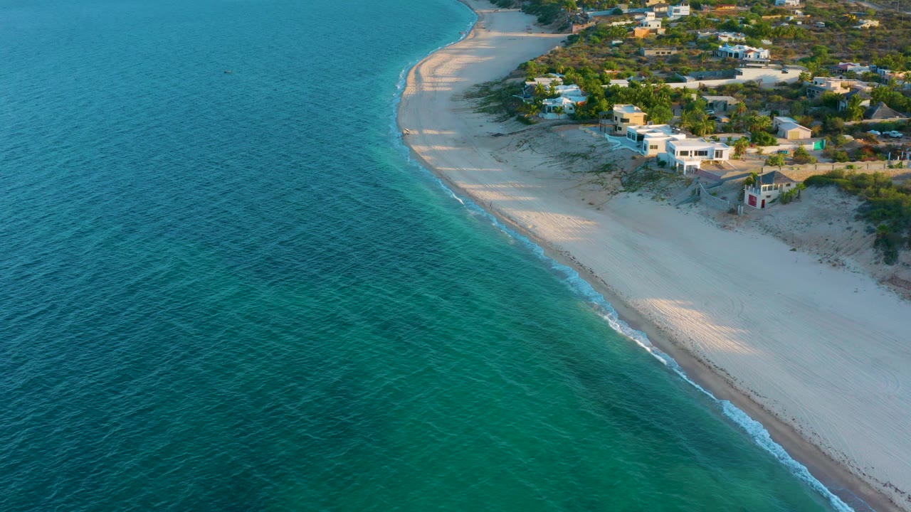 Aerial coastal frame in El Sargento La Ventana, Mexico with clear blue-green water across the lower half, a pale beach curve, scattered homes, trees, dunes, and no visible people.