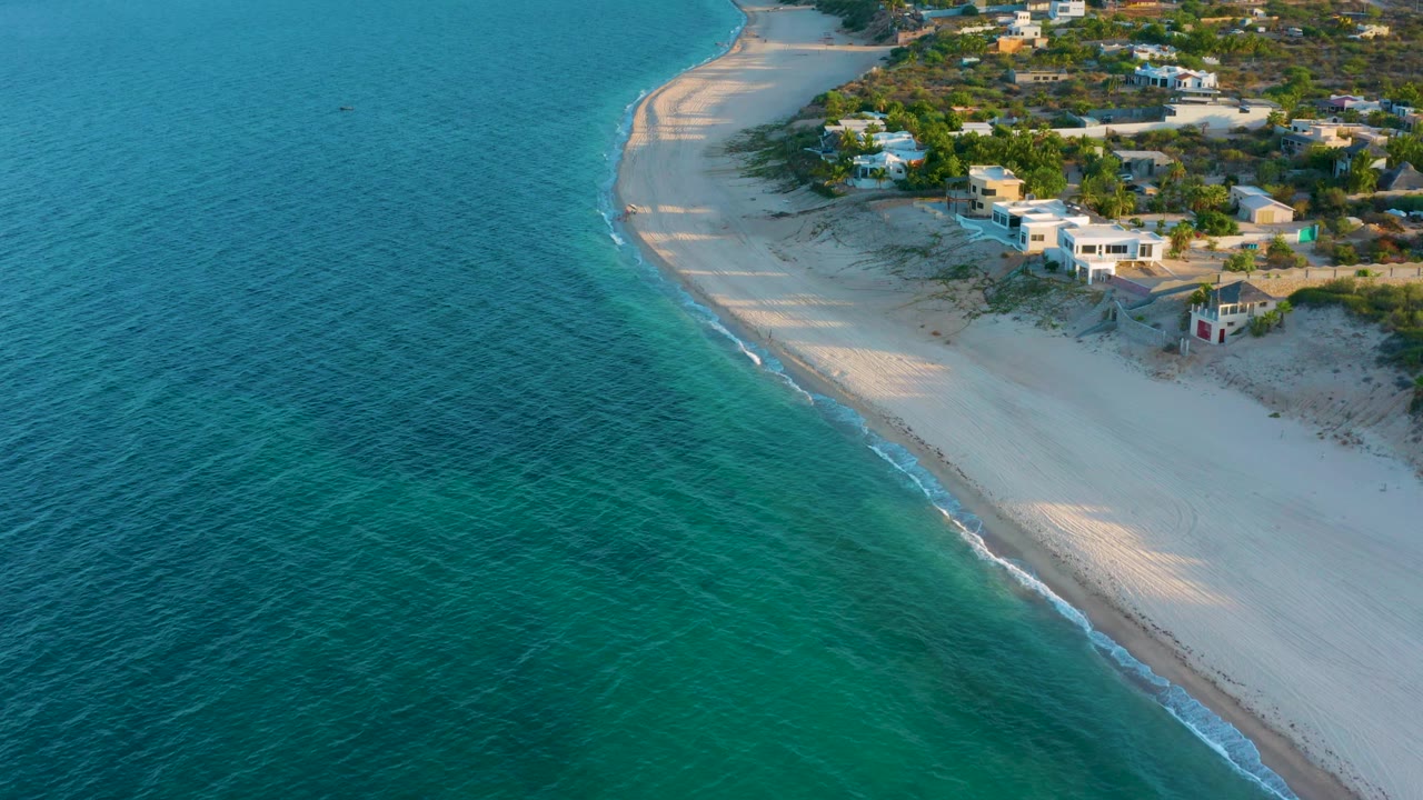 Drone view of sandy coastline with homes, trees, and calm surf.