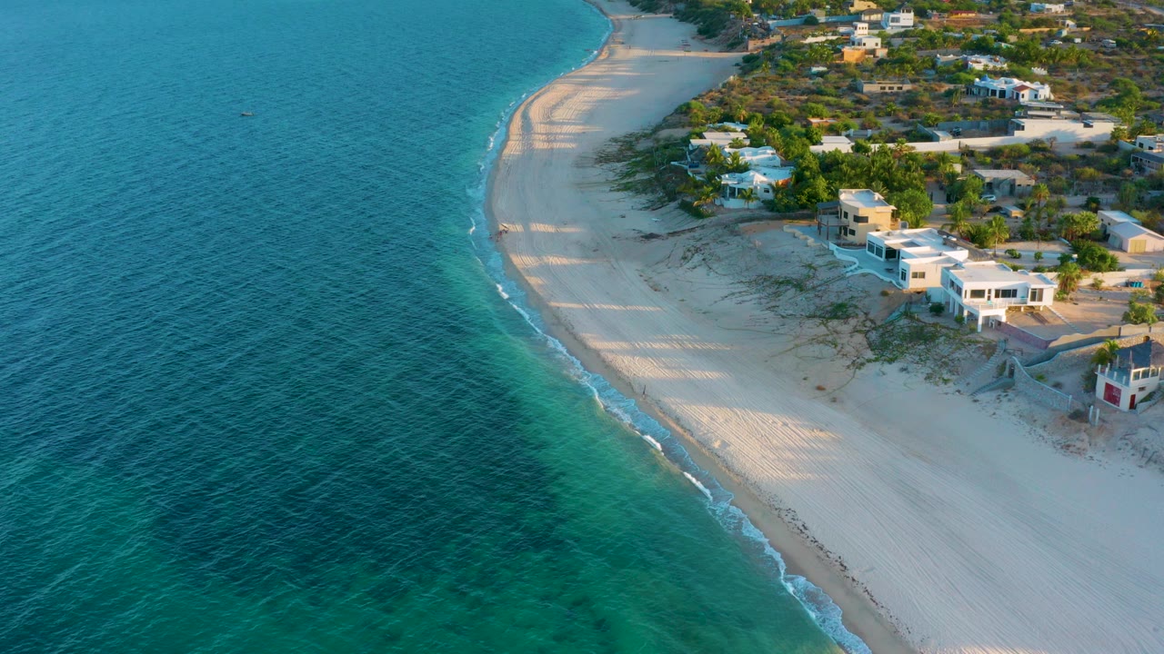 Opening aerial frame over El Sargento La Ventana, Mexico, with turquoise water at left, a curved pale beach, low homes by the sand, and no visible people.