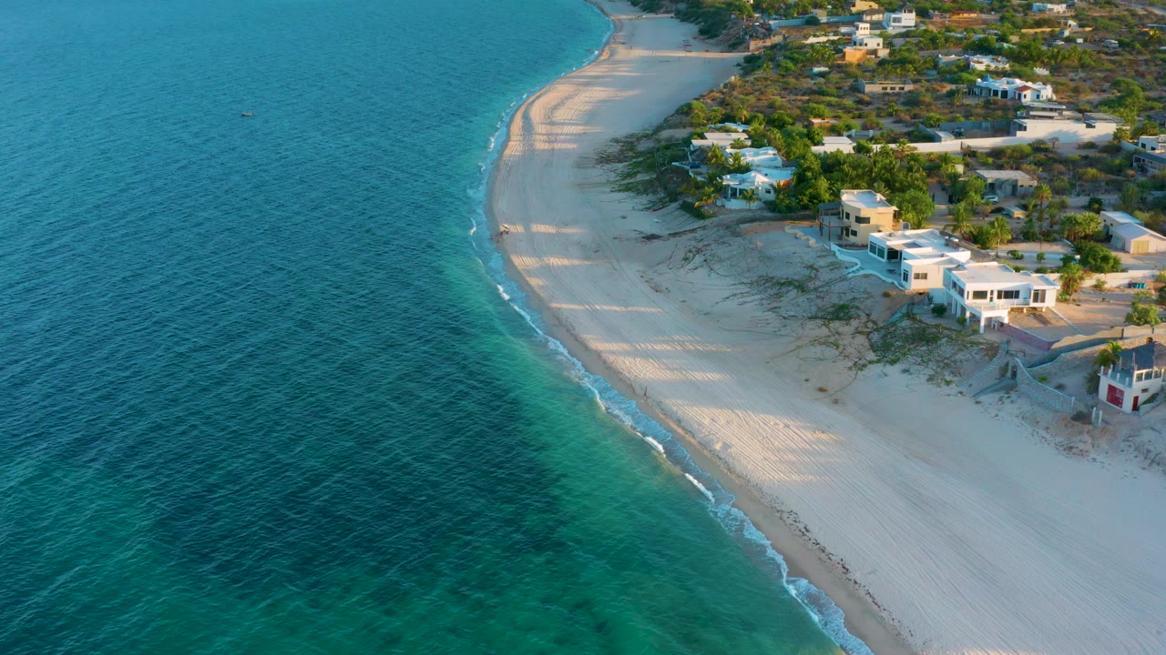 Aerial view of El Sargento La Ventana, Mexico, showing more open blue water on the left, white sand curving upward, beach houses on the right, and no visible people.