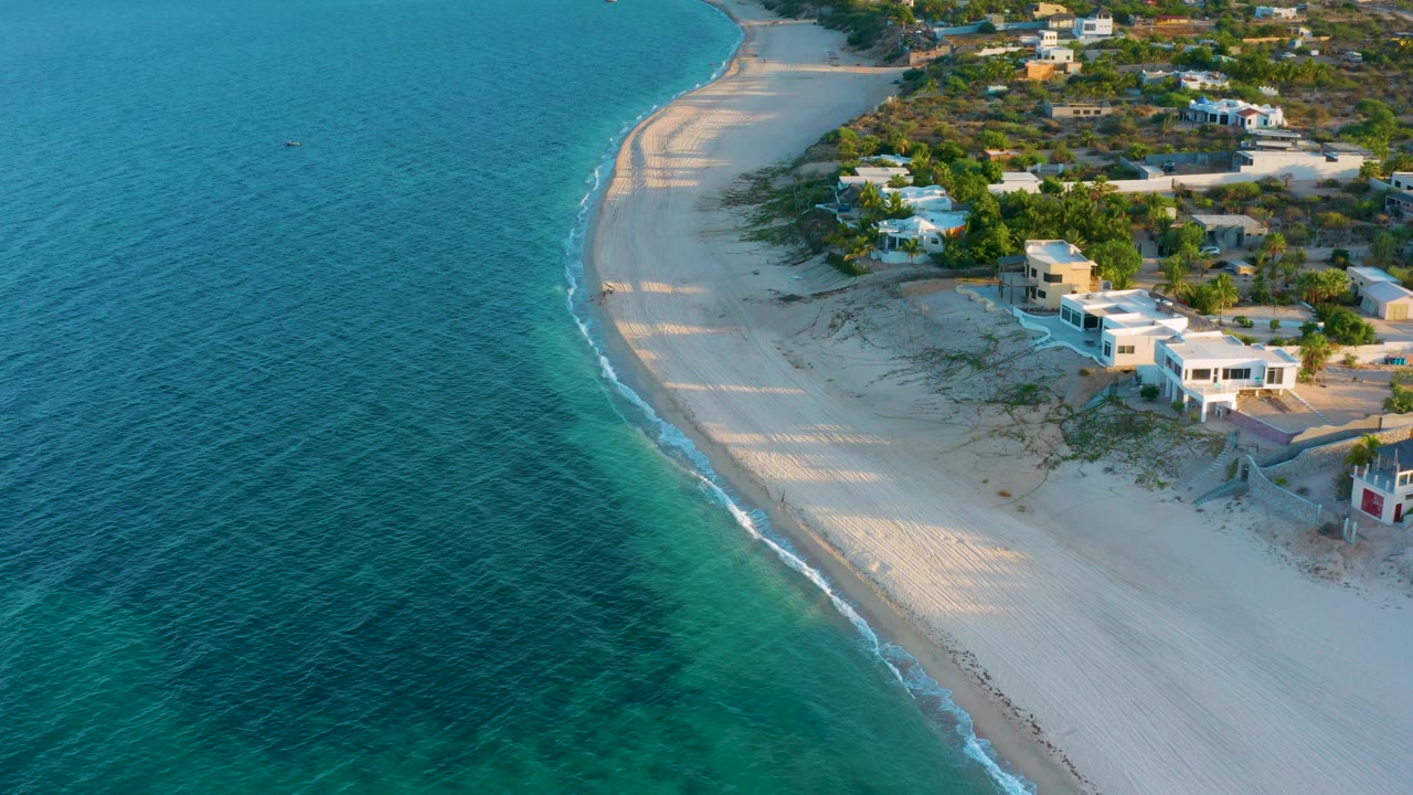 Aerial frame over El Sargento La Ventana, Mexico, with teal shallows, a narrow surf line, bright sand, low homes near the upper right, and no visible people.