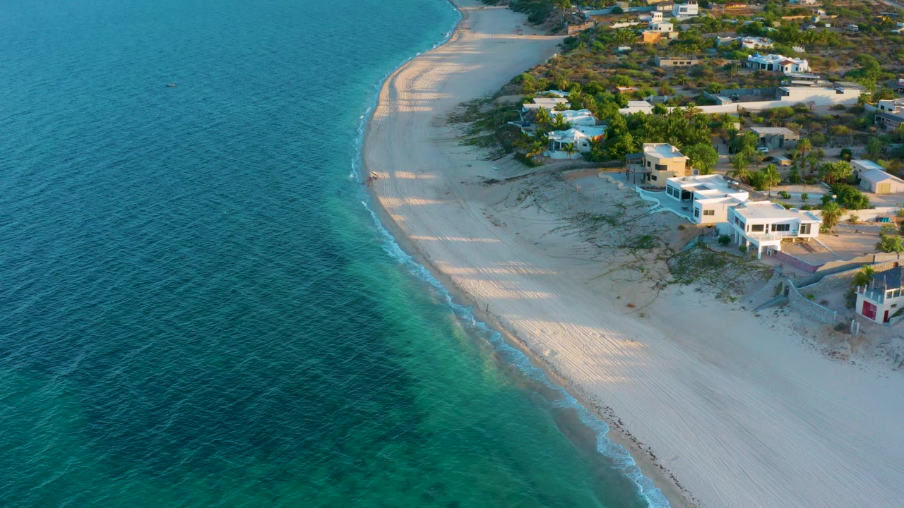 High aerial frame over El Sargento La Ventana, Mexico showing blue-green water, a narrow line of surf, bright sand, shoreline houses, and no visible people.