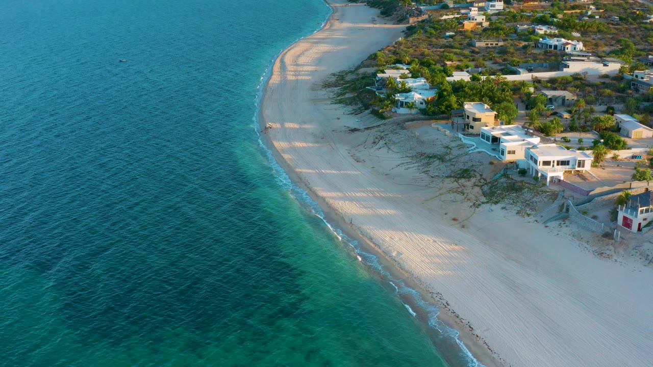 Aerial view in El Sargento La Ventana, Mexico with teal ocean filling the left side, a smooth sandy beach through the center, low homes and brush to the right, and no visible people.