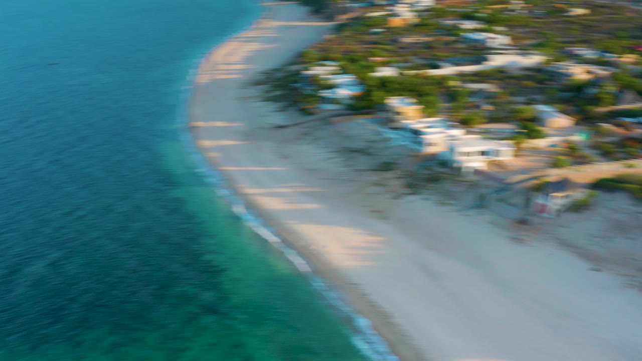 High aerial frame over El Sargento La Ventana, Mexico, with blue-green water at left, pale sand along the shore, blurred coastal homes beyond, and no visible people.