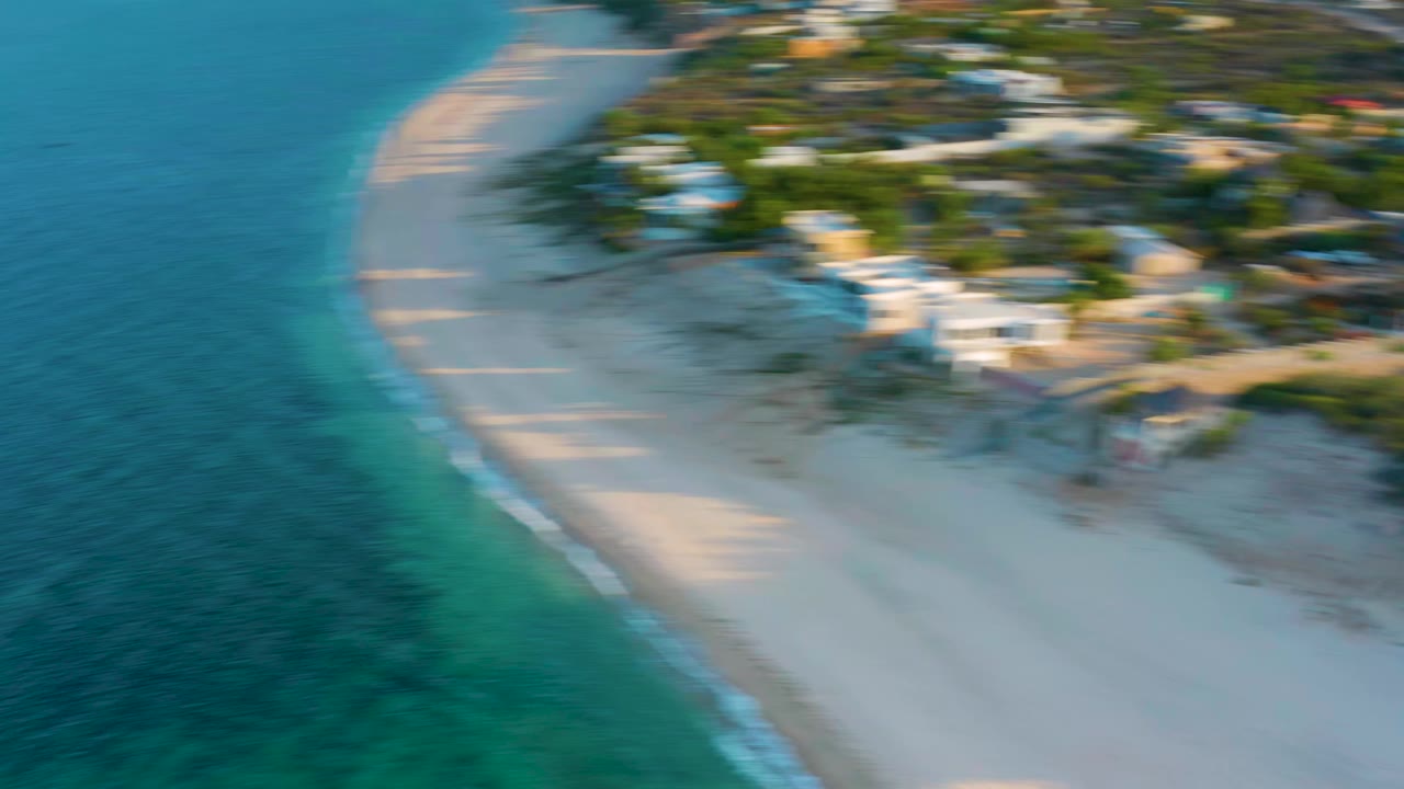 Aerial view in El Sargento La Ventana, Mexico, showing the curving beach, clear water on the left, softened inland houses from motion blur, and no visible people.