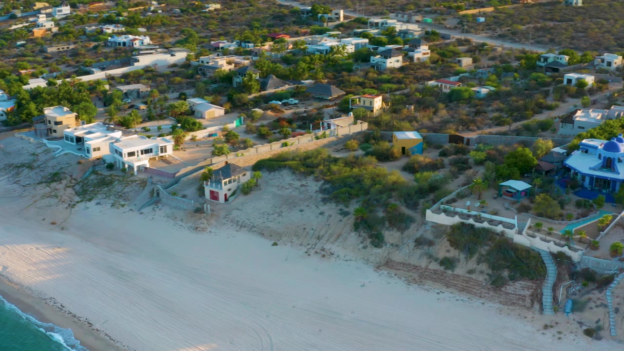 A clearer aerial frame over El Sargento La Ventana, Mexico, with beachfront houses, sandy dunes, pale beach, turquoise water at the lower edge, and no visible people.