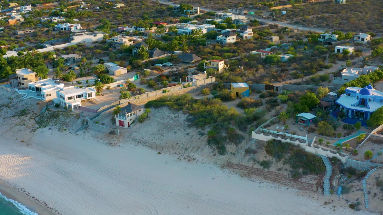 Aerial view of coastal homes, scrubland, sand, and water at the edge of frame.