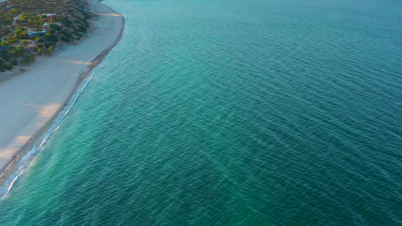 Opening aerial frame over El Sargento La Ventana, Mexico, with pale sand at left, blue-green water across the frame, shoreline trees and structures near the top left, and no visible people.