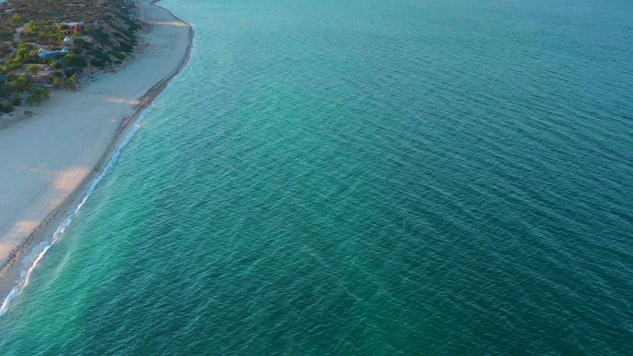 Aerial view over El Sargento La Ventana, Mexico, with a narrow sand strip at left, small white wave lines along the shore, rippled turquoise water extending right, and no visible people.