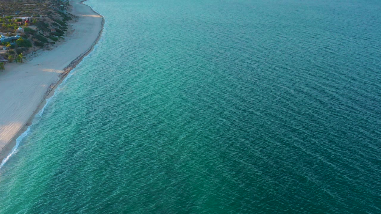 Aerial view in El Sargento La Ventana, Mexico, showing rippled blue green sea, a narrow pale beach at left, a thin line of surf, and no visible people.