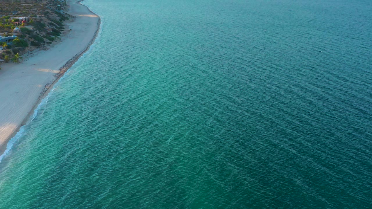 The frame shows El Sargento La Ventana coastline from above, with clear blue water on the right, pale beach and trees on the left, and no visible people.