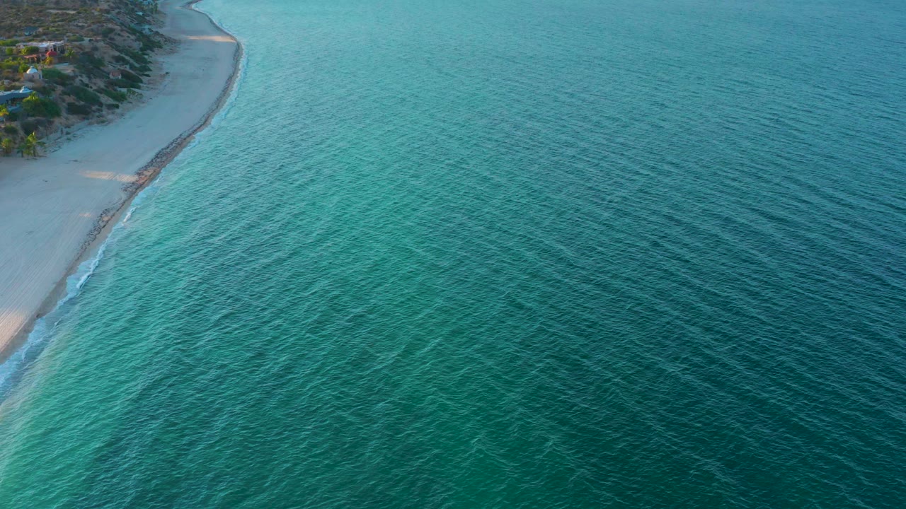 Aerial frame from El Sargento La Ventana, Mexico, with shallow turquoise water crossing the foreground, an empty pale beach at left, and no visible people.
