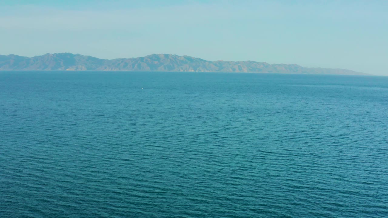 Open blue ocean with subtle texture leads to hazy distant mountains and a pale sky near El Sargento La Ventana, Mexico, with no visible people.