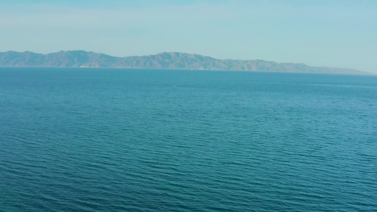 Open blue water stretches toward hazy mountains under a clear pale sky near El Sargento La Ventana, Mexico, with no boats or people visible.