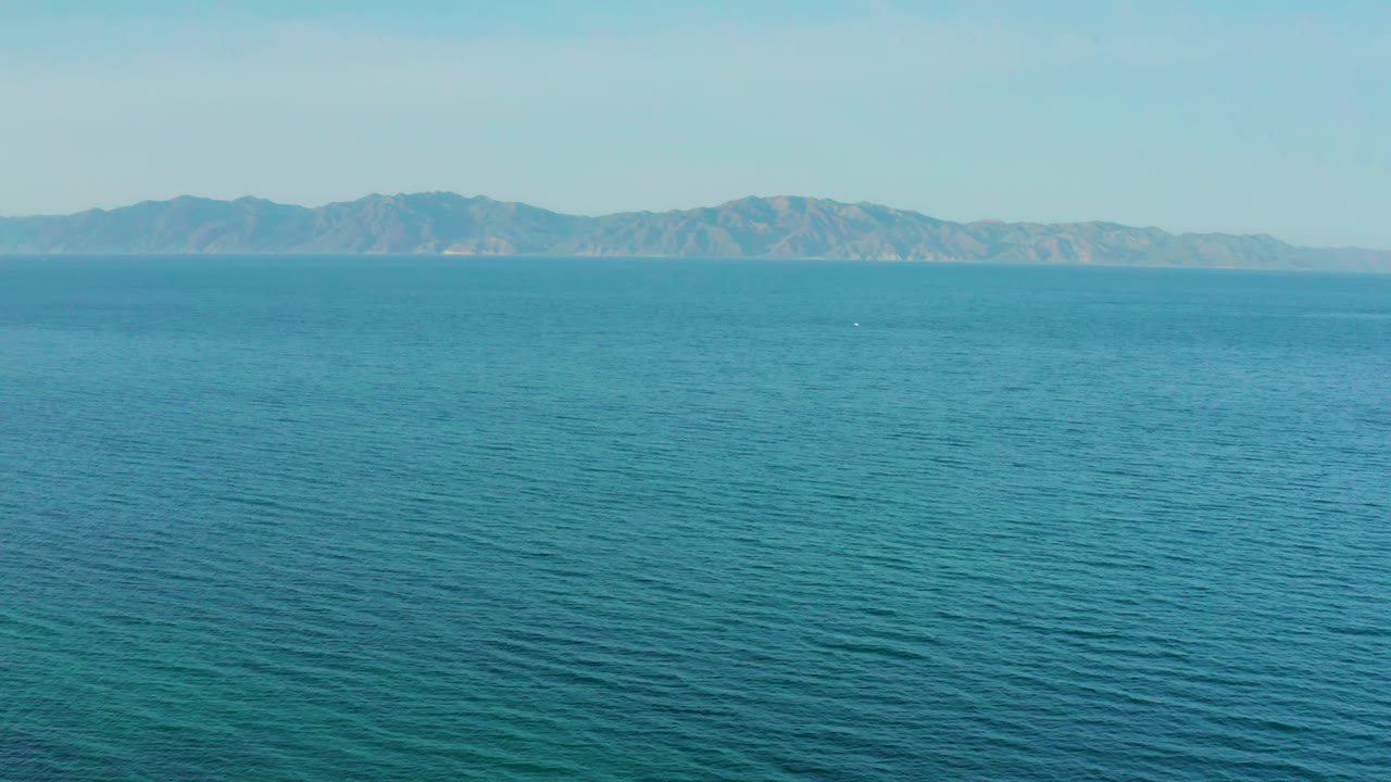 Calm turquoise water fills the foreground while hazy mountains stretch across the horizon near El Sargento La Ventana, Mexico, with no visible people.