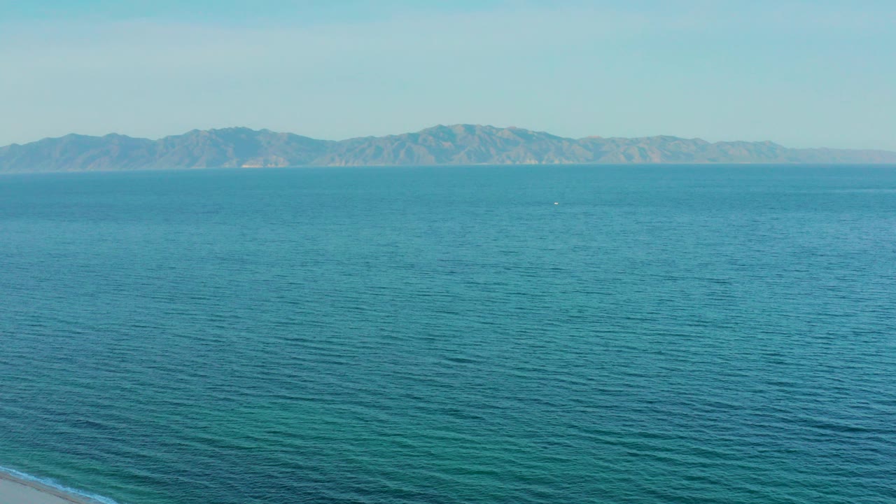 Wide aerial frame over blue-green water near El Sargento La Ventana, Mexico, with a thin strip of beach at the bottom edge, distant low mountains on the horizon, and no visible people.