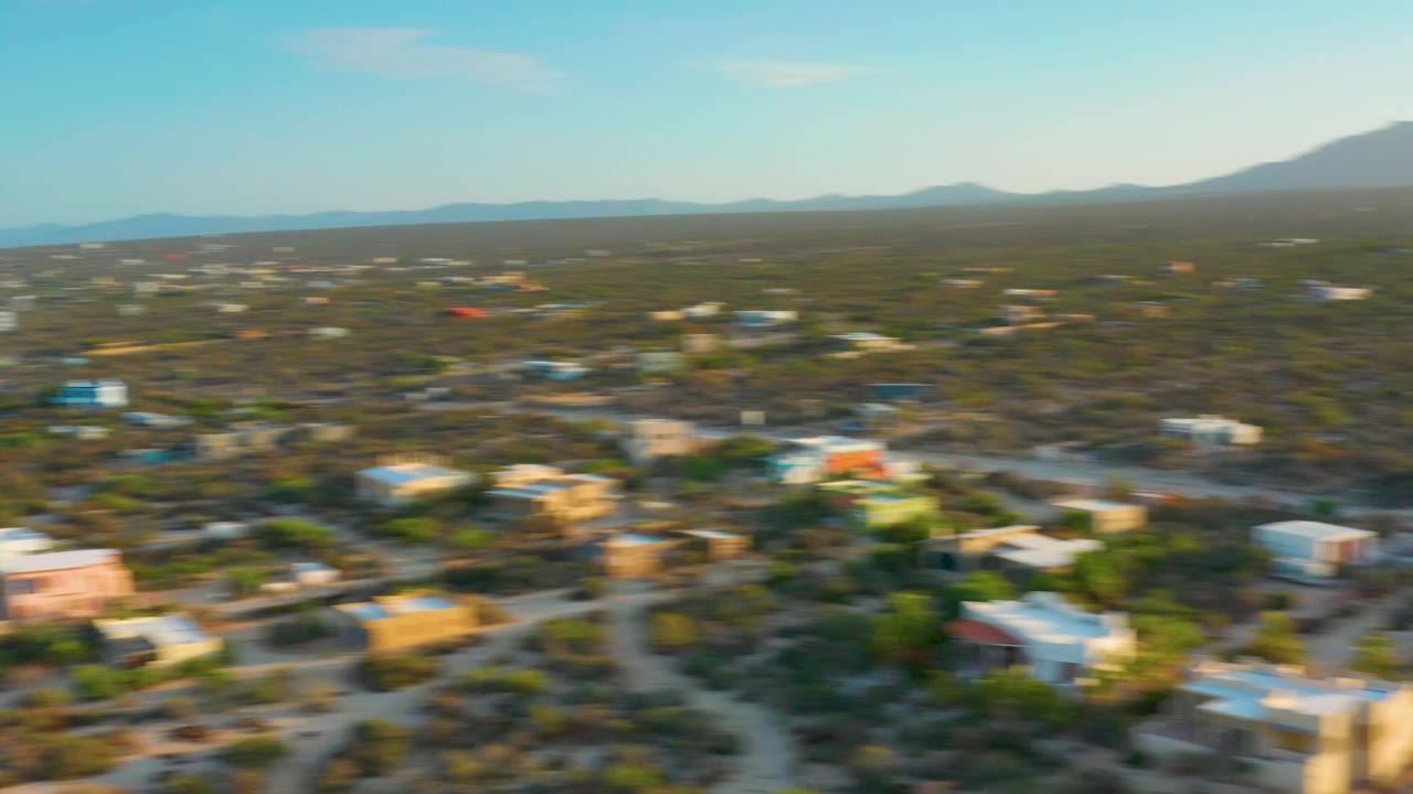 Aerial frame over El Sargento La Ventana with visible motion blur across houses and desert shrubs, mountains in back, blue sky above, and no visible people.