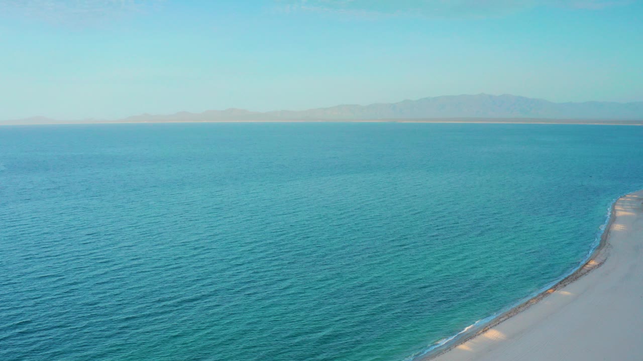 Aerial frame from El Sargento La Ventana, Mexico showing turquoise water, a pale shoreline entering from the lower right, distant mountains, and no visible people.