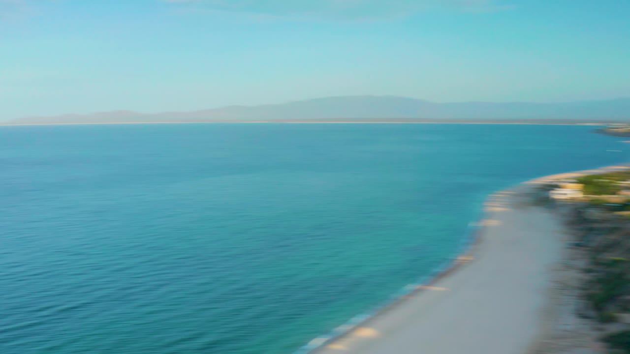Aerial coastal frame from El Sargento La Ventana, Mexico showing open turquoise water, sandy beach along the right edge, a faint mountain horizon, and no visible people.