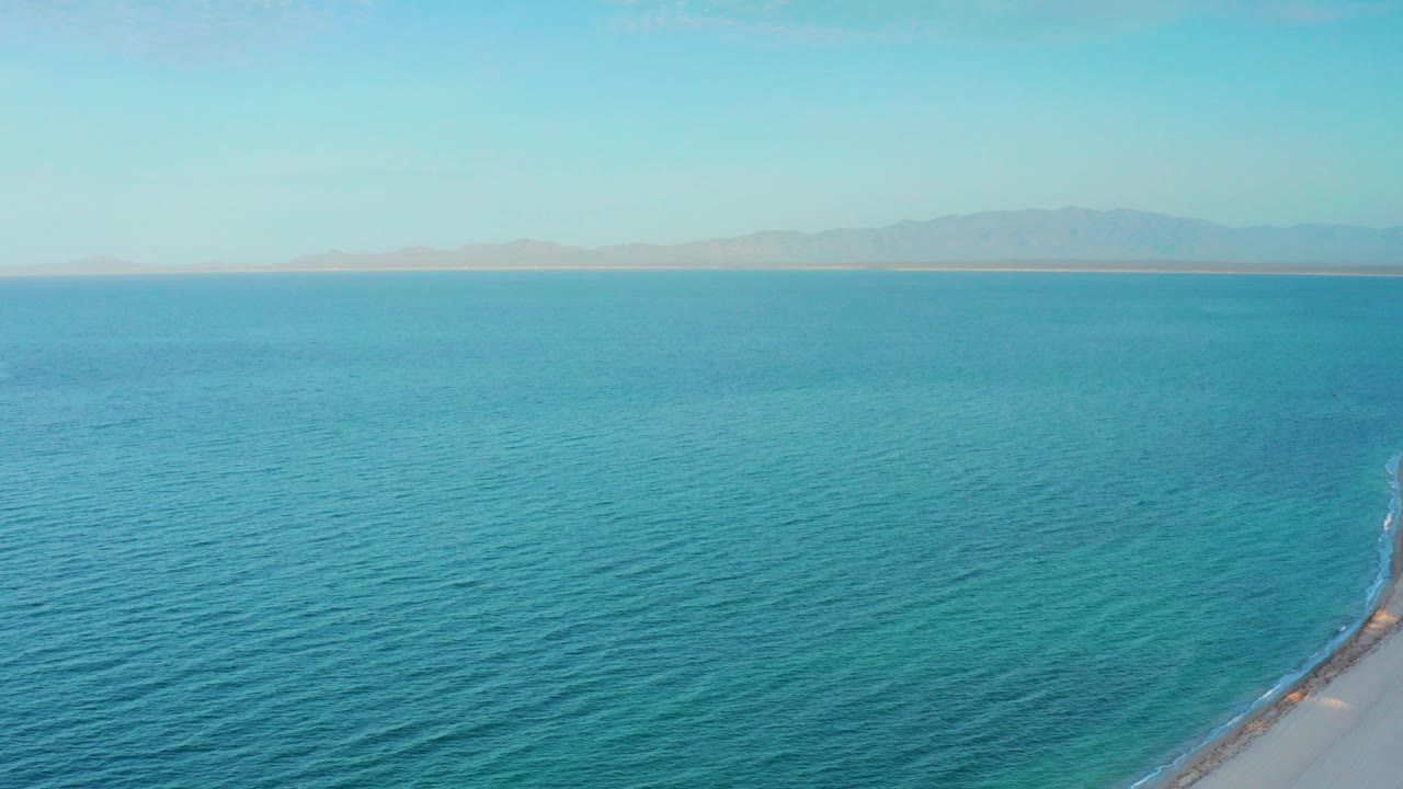 Aerial view over blue ocean at El Sargento La Ventana, Mexico, with distant hazy mountains, a pale sky, a small shoreline point at lower right, and no visible people.