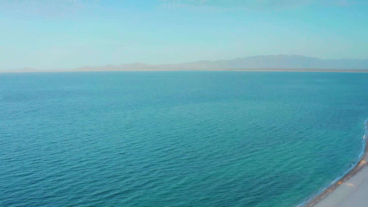 Wide aerial view at El Sargento La Ventana, Mexico, showing turquoise ocean, a hazy mountain horizon, faint sandy shore at lower right, and no visible people.