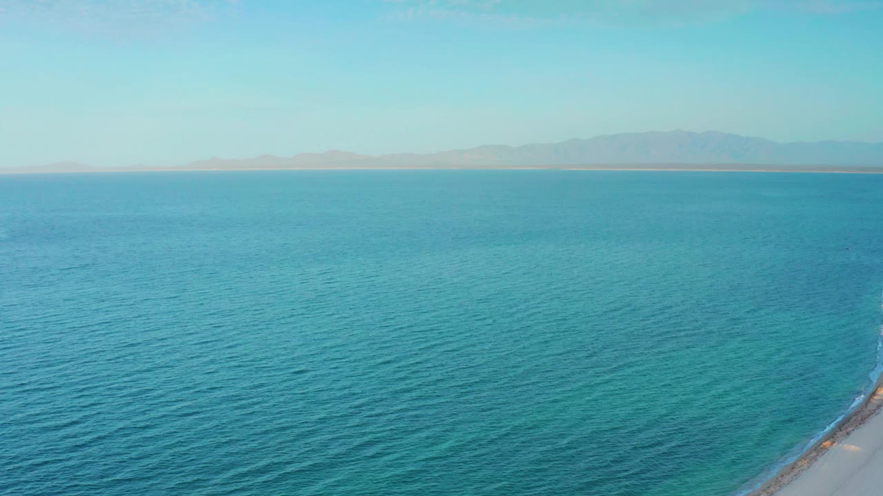 High aerial frame from El Sargento La Ventana, Mexico, with calm blue water across the foreground, distant mountains under a pale sky, a narrow shoreline strip at right, and no visible people.