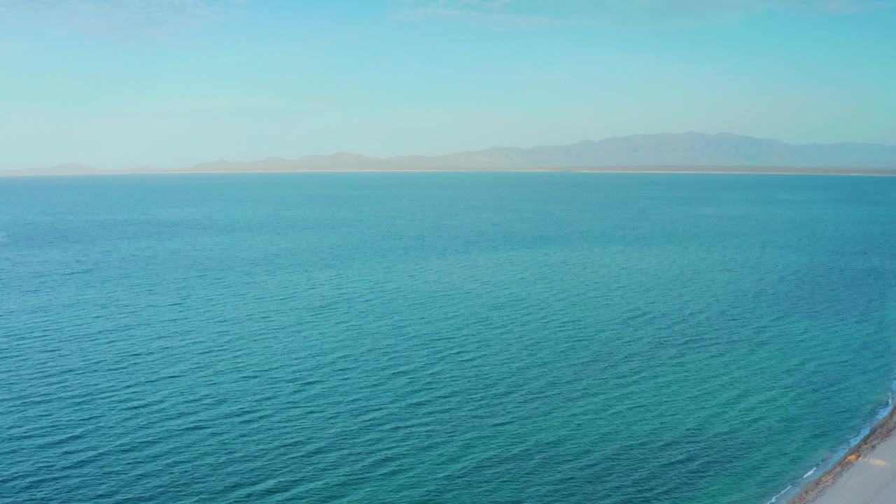 Aerial ocean frame at El Sargento La Ventana, Mexico, with textured blue water, a small rocky shoreline point near the lower right corner, hazy mountains, and no visible people.
