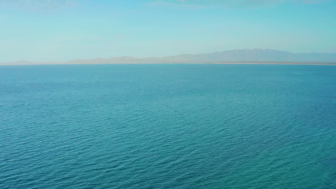 Wide view of blue water off El Sargento La Ventana, Mexico, with a thin distant shore and hazy mountains under clear sky, no visible people.