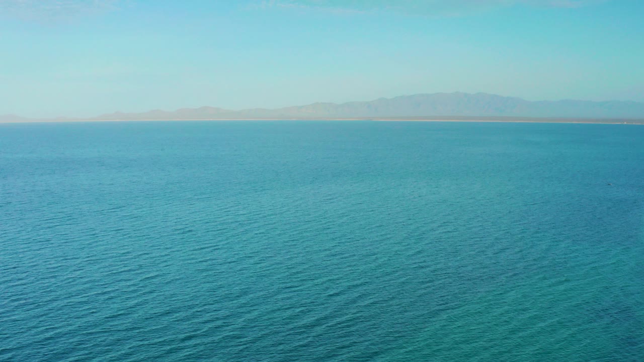 Wide aerial view over blue ocean near El Sargento La Ventana, Mexico, with faint landforms across the horizon and no visible people.