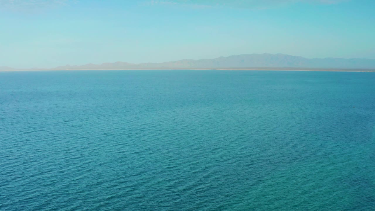 Open ocean fills the frame with a pale sky above and a distant shoreline running across the horizon near El Sargento La Ventana, Mexico, with no visible people.
