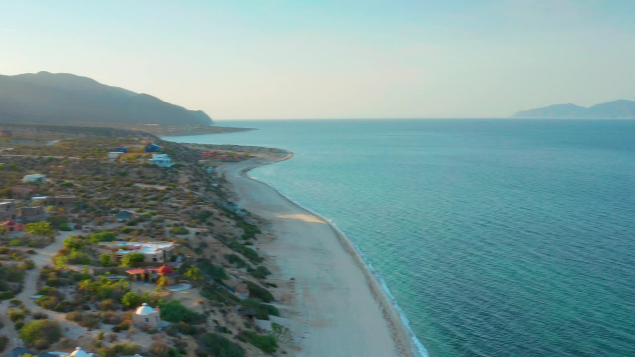 Opening aerial frame over El Sargento La Ventana, Mexico, showing a pale beach curving beside turquoise rippled water, scattered coastal buildings, scrub vegetation, distant mountains, and no visible people.