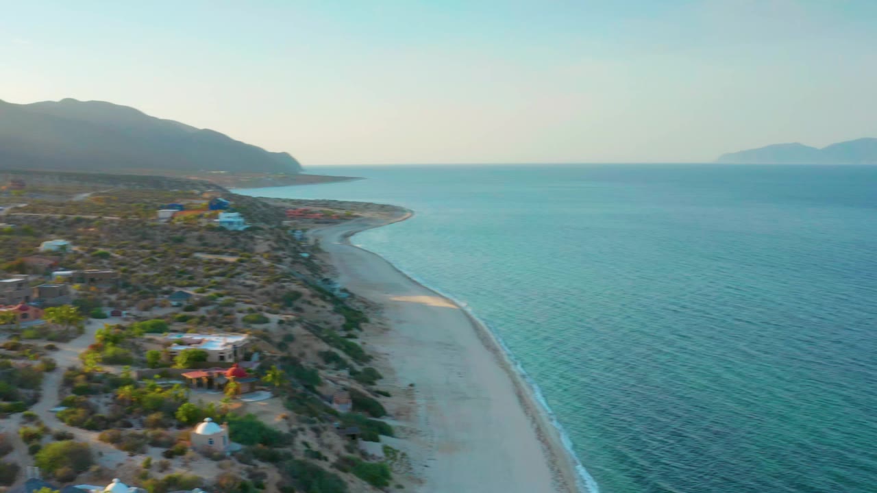 One second into the clip, the aerial view holds on El Sargento La Ventana coastline with more open water on the right, scattered small buildings on the left, mountains beyond the shore, and no visible people.