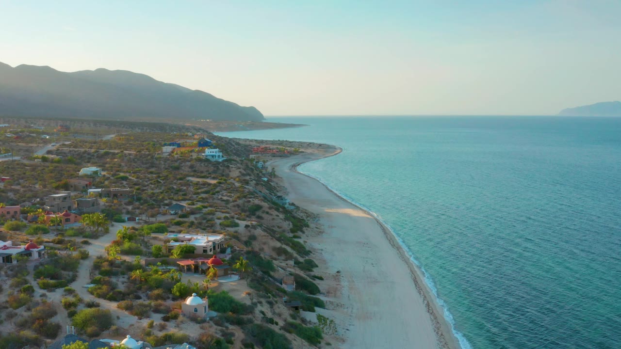 At 0:02, the El Sargento La Ventana shoreline bends through frame, with turquoise water textured by wind, small houses and vegetation inland, hazy mountains at left, and no visible people.