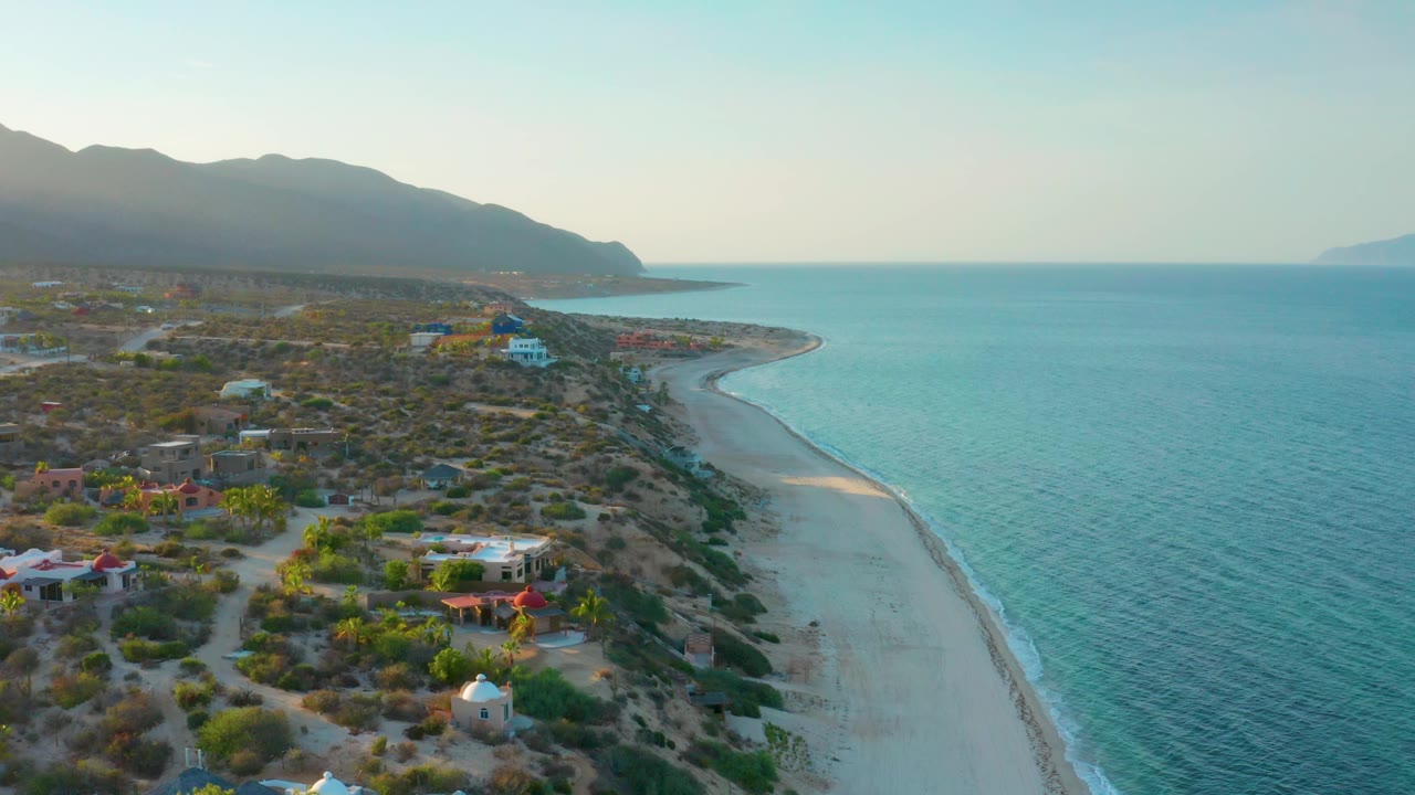 Wide aerial shoreline with beach, sea, scattered homes, scrub, and mountains.
